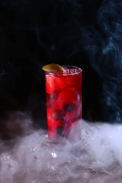 Homemade lemonade made from berries in a tall glass. Vertical photo on a black background. Chunks of ice. Smoke on a black background. — Stock Photo, Image