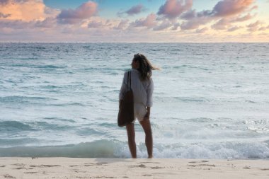 Sahilde, bluz ve spor çantası giyen bir kadın Meksika 'daki Karayip Denizi' nin üzerindeki ufka bakıyor. Arka planda gün batımında gökyüzü