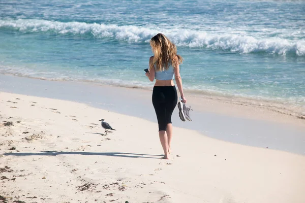 Tanınamayacak kadar sevimli bir kadın tropik bir plajın beyaz kumlarında yalın ayak yürür, akıllı telefonuna bakar. Arka planda Meksika Karayip Denizi