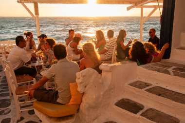 A group of people sitting in a bar in the village on the island of Mykonos in Greece on the waterfront at sunset. In the background the bright sky and the Mediterranean sea