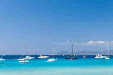 Formentera adasındaki bir plajın yakınındaki turkuaz denizde demirlemiş çeşitli yelkenliler. Arka planda mavi gökyüzü ve İbiza kıyıları