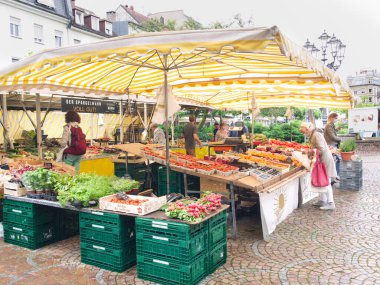 Sebze, meyve ve otların sergilendiği bir sokak pazarında oyalamak. Siyahi maskeli insanlar bakkaldan alışveriş yapıyorlar. Almanya 'nın Baden-Baden kasabası.