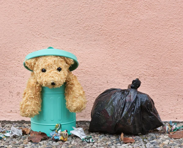 A soft toy representing an abandoned dog dumped in a trash can. A garbage bag and other litter nearby.