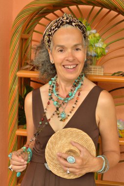 Kahverengi yaz kıyafetleri ve bir sürü turkuaz mücevher takan güler yüzlü zarif bir kadın. Arka planda egzotik bir kitaplık.