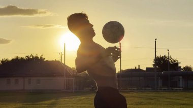 Genç adam gün batımında stadyumda futbol topuyla hokkabazlık yapıyor. Yeşil sahada profesyonel futbolcu topa vuruyor. Sporcu arka planda güneş ışığıyla çayırda numaralar yapıyor. Serbest stil futbol.