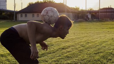 Genç adam gün batımında stadyumda futbol topuyla hokkabazlık yapıyor. Yeşil sahada profesyonel futbolcu topa vuruyor. Sporcu arka planda güneş ışığıyla çayırda numaralar yapıyor. Serbest stil futbol.
