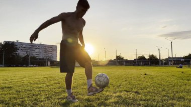 Genç adam gün batımında stadyumda futbol topuyla hokkabazlık yapıyor. Yeşil sahada profesyonel futbolcu topa vuruyor. Sporcu arka planda güneş ışığıyla çayırda numaralar yapıyor. Serbest stil futbol.
