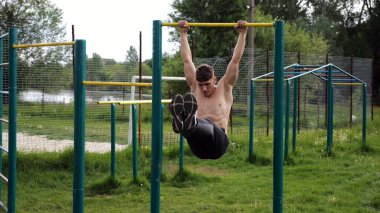 Atletik adam karın kaslarını eğitir spor sahasında yatay barda bacak kaldırır. Genç sporcu, barfiks çekme çubuğunda egzersiz yapıyor. Doğada spor yapan sporcu. Sağlıklı yaşam tarzı.