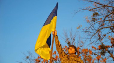 Erkek Ukraynalı asker, Rus saldırısına karşı zafer şerefine mavi-sarı bayrak kaldırıyor. Kamuflaj üniformalı genç ordu Ukrayna bayrağını kırsala çekti. İstila direnişi.