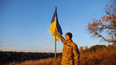 Üniformalı erkek asker, gün batımında Rus saldırısına karşı kazanılan zaferin şerefine mavi-sarı bayrak kaldırıyor. Ukrayna ordusunun genç askeri Ukrayna bayrağını taşraya salladı..