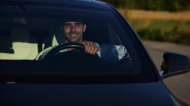 Modern elektrikli arabanın ön camından bakan yakışıklı başarılı bir girişimci. Yenilikçi elektrikli aracında gülümseyen genç iş adamının portresi. Yavaş çekim
