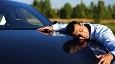 Successful smiling manager bought new electrical vehicle and hugging hood of auto. Happy young businessman in shirt enjoying modern electric automobile. Concept of buy or rent car. Slow mo
