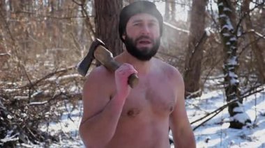 Tişörtsüz sakallı adamın portresi karlı bir kış ormanında baltayla duruyor. Karlı ormanlık alanda odun kestikten sonra kameraya bakan yorgun bir adam. Sahne, soğuk koşullarda açık havada çalışmayı temsil ediyor..