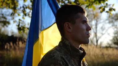 Ukraynalı asker kırsal kesimde ulusal pankartıyla ayakta duruyor. Rus saldırısına karşı zafer sembolü olarak Ukrayna bayrağı taşıyan askeri üniformalı bir erkek asker. İstila direnişi kavramı.