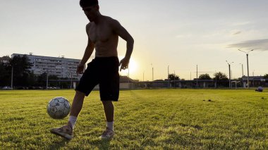 Genç adam gün batımında stadyumda futbol topuyla hokkabazlık yapıyor. Yeşil sahada profesyonel futbolcu topa vuruyor. Sporcu arka planda güneş ışığıyla çayırda numaralar yapıyor. Serbest stil futbol.