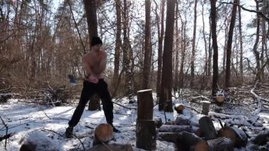 Güneşli bir kış ormanında yakacak odun kesen üstsüz genç bir adam. Karlı ormanlık alanda baltayla çalışan kaslı bir adam. Güç kavramı, erkeksi, enerji ve soğuk mevsim boyunca doğayla uyum..