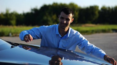 Successful smiling manager bought new electrical vehicle and hugging hood of auto. Happy young businessman in shirt enjoying modern electric automobile. Concept of buy or rent car. Slow mo