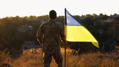 Ukrayna ordusunun askeri gün batımında mavi-sarı pankartı kaldıracak. Üniformalı genç erkek asker Ukrayna bayrağıyla kırsalda yürüyor. Rus saldırganlığına karşı zafer.