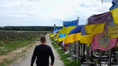 Mourning man go among countless graves of ukrainian soldiers with national flags at military cemetery in Kharkiv. Scene reflects personal sorrow and nation collective loss caused by russian aggression.