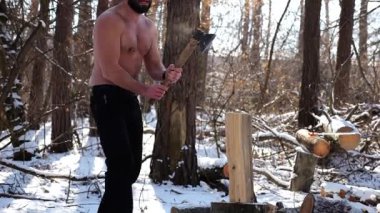 Güneşli kış ormanlarında odun kesen kaslı sakallı adam. Karlı ormanlık alanda baltayla çalışan üstsüz genç bir adam. Güç kavramı, erkeksi, enerji ve soğuk mevsim boyunca doğayla uyum..