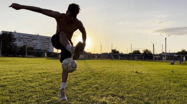 Genç adam gün batımında stadyumda futbol topuyla hokkabazlık yapıyor. Yeşil sahada profesyonel futbolcu topa vuruyor. Sporcu arka planda güneş ışığıyla çayırda numaralar yapıyor. Serbest stil futbol.