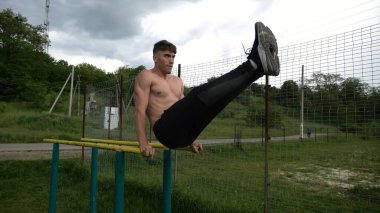 Güçlü bir adam karın kaslarını eğitir spor sahasında paralel barlarda bacak kaldırır. Genç üstsüz sporcu doğada egzersiz yapıyor. Doğada spor yapan sporcu. Sportif yaşam tarzı kavramı.