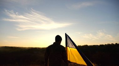 Ukraynalı askerin silueti gün batımında kırsal alanda ulusal bayrağın önünde duruyor. Rus saldırısına karşı zafer sembolü olarak Ukrayna bayrağı taşıyan askeri üniformalı bir erkek asker. Yavaş çekim