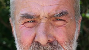 Yaşlı bir adamın mavi gözleri yanıp sönüyor ve dışarıdaki kameraya bakıyor. Üzücü bir manzarası olan yaşlı bir insanın buruşuk yüzü. Emeklinin üzgün yüz ifadesi.