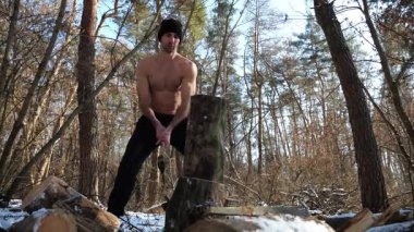 Güneşli bir kış ormanında yakacak odun kesen üstsüz genç bir adam. Karlı ormanlık alanda baltayla çalışan kaslı bir adam. Güç kavramı, erkeksi, enerji ve soğuk mevsim boyunca doğayla uyum..