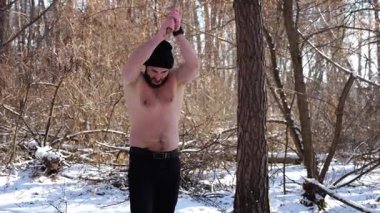 Gömleksiz kaslı adam, karlı ormanda baltasını dikiyor. Güçlü adam, dayanıklılık, güç ve güven göstererek kaslarını esnetir. Sahne, açık hava aktivitesi ve gücü harmanlıyor..