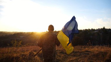 Askeri üniformalı genç adam gün batımında Ukrayna bayrağı sallayarak koşuyor. Ukrayna ordusunun erkek askeri kırsalda mavi-sarı pankartla koşuyordu. İstila direnişi kavramı.