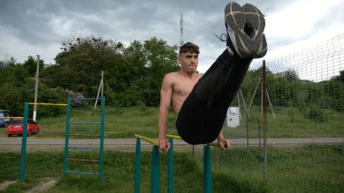 Güçlü bir adam karın kaslarını eğitir spor sahasında paralel barlarda bacak kaldırır. Genç üstsüz sporcu doğada egzersiz yapıyor. Doğada spor yapan sporcu. Sportif yaşam tarzı kavramı.