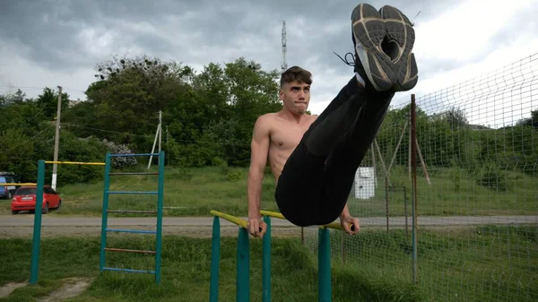 Güçlü bir adam karın kaslarını eğitir spor sahasında paralel barlarda bacak kaldırır. Genç üstsüz sporcu doğada egzersiz yapıyor. Doğada spor yapan sporcu. Sportif yaşam tarzı kavramı.