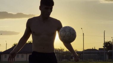 Genç adam gün batımında stadyumda futbol topuyla hokkabazlık yapıyor. Yeşil sahada profesyonel futbolcu topa vuruyor. Sporcu arka planda güneş ışığıyla çayırda numaralar yapıyor. Serbest stil futbol.