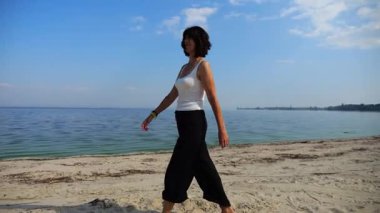 Güneşli bir günde sahilde kumsala giden mutlu yetişkin kadın. Arka planda sakin dalgalarla deniz kıyısında yürüyen güzel bir bayan. Yaz tatili ya da tatil kavramı. Yavaş çekim