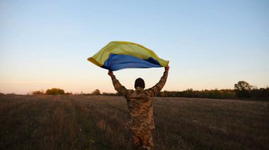 Ukrayna bayrağını dalgalandıran Ukrayna ordusunun askeri. Genç erkek asker üniformalı, çayırda mavi-sarı pankartla koşuyor. Rus saldırısına karşı zafer.