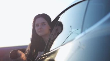 Genç, güzel bir kız, modern elektrikli arabaya doğru eğilmiş ve kameraya bakıyor. Alternatif elektrikli araçta gülümseyen çekici bir kadının portresi. Yavaş çekim