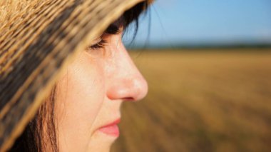 Hasır şapkalı kadın çiftçi buğday tarlasına bakıyor ve gün batımında çiftliği inceliyor. Güneşin parlak ışıklarıyla aydınlatan yetişkin ve güzel bir kadının yüzünü kapatın. Tarım sektörü kavramı.