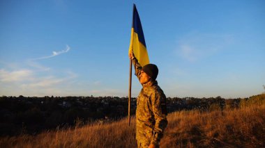 Üniformalı erkek asker, gün batımında Rus saldırısına karşı kazanılan zaferin şerefine mavi-sarı bayrak kaldırıyor. Ukrayna ordusunun genç askeri Ukrayna bayrağını taşraya salladı..