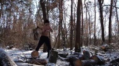 Güneşli bir kış ormanında yakacak odun kesen üstsüz genç bir adam. Karlı ormanlık alanda baltayla çalışan kaslı bir adam. Güç kavramı, erkeksi, enerji ve soğuk mevsim boyunca doğayla uyum..