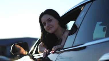 Genç, güzel bir kız, modern elektrikli arabaya doğru eğilmiş ve kameraya bakıyor. Alternatif elektrikli araçta gülümseyen çekici bir kadının portresi. Yavaş çekim