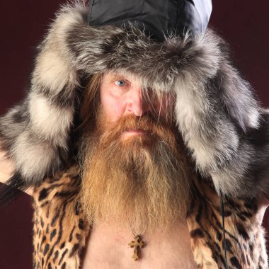 Uzun sakallı ve bıyıklı bir adamın yakın plan portresi, kürk şapkalı, bordo bir arka planda stüdyo fotoğrafı.