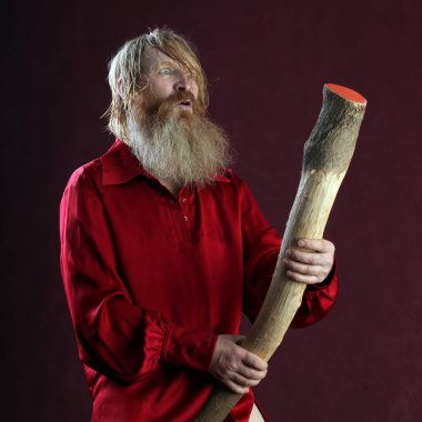 Kırmızı gömlekli, uzun sakallı ve bıyıklı bir adamın portresi. Elinde copu ve bordo bir arka planda el yapımı ayakkabıları var.