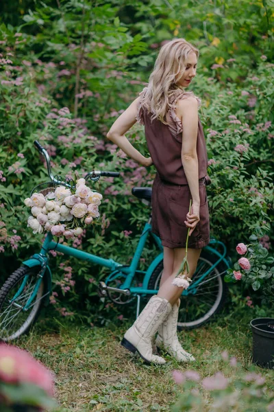 Uzun sarı saçlı güzel bir kız eski bir bisikletin yanında dikilirken pembe şakayık çiçeği tutuyor.
