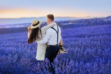 Guy carries happy girlfriend in a white dress by his arms at beautiful sunset time at purple lavender field. Airy and peaceful horizontal photo taken during trip to Provence, France