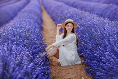 Beautiful smiling  brunette girl in white vintage dress and straw hat sits in a lavender field with a dreamy mood and looks to the left