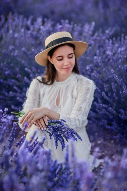 Emotional photo  of beautiful smiling  brunette girl in white vintage dress and straw hat sits in a lavender field with a dreamy mood closing her eyes. Vertical photo