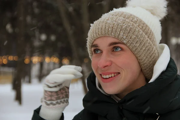 Smiling blue-eyed girl in a knitted hat raised her hand in knitted gloves against the background of a winter park