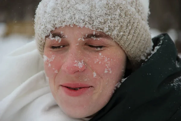 Face of a smiling girl with closed eyes in snowflakes close-up in a knitted hat