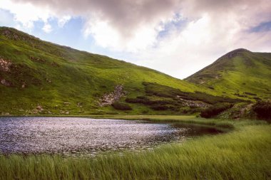 Dağların üzerinde bulutlu bir gökyüzü, dağlık bir göl. Suyun üzerinde dalgalar ve çimlerin etrafında dalgalar.